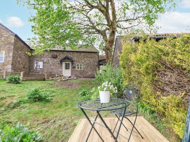 A backyard with a stone building a tree a metal table and chair with flowers on the table at Cuddfan at Swallow Cottage in Rhydtalog near Coedpoeth