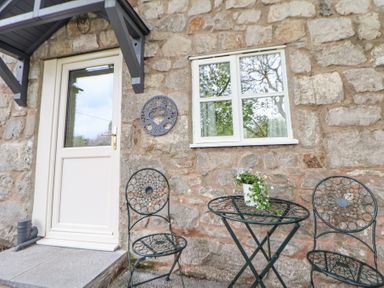 An outdoor seating area with two metal chairs and a metal table with a flower pot in front of a stone wall with a door and window at Cuddfan at Swallow Cottage in Rhydtalog near Coedpoeth