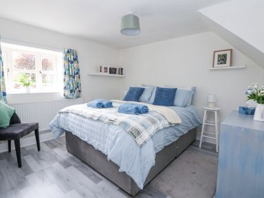 A bedroom with a double bed pillows towels and bedding a window with patterned curtains a chair and shelves with books and a framed photo at Cuddfan at Swallow Cottage in Rhydtalog near Coedpoeth