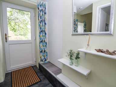 An entryway with a white door a mat patterned curtains a mirror and two shelves with glass vases and decorative items at Cuddfan at Swallow Cottage Rhydtalog near Coedpoeth