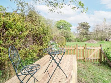 A wooden deck with a metal table and two chairs next to a wooden gate and green bushes at Cuddfan at Swallow Cottage in Rhydtalog near Coedpoeth