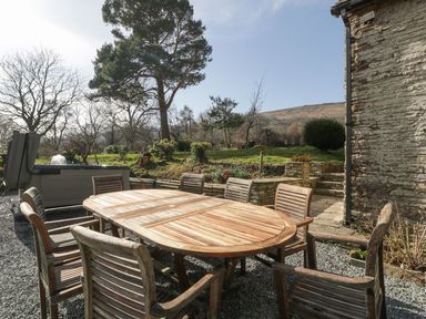 An outdoor wooden dining table with chairs on a gravel patio next to a stone wall with trees and a hill in the background at White Haywood Farmhouse & Barn in Craswall