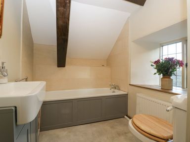 A bathroom with a bathtub under a sloped ceiling a sink a toilet with a wooden seat and a window with flowers on the sill at White Haywood Farmhouse & Barn Craswall