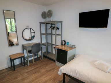 A bedroom with a bed a wall mounted tv a small desk and chair a stool an open wardrobe with hangers two fans on top and a mini fridge at Beech in Cowbridge