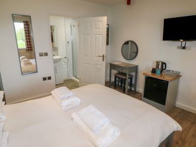 A bedroom with a bed and folded towels on it a wall mirror a desk with a round mirror a mini fridge with a kettle and mugs and a wall-mounted television at Cherry in Cowbridge