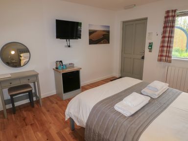 A bedroom with a bed with folded towels a small desk with a stool a wall-mounted TV a kettle on a cabinet and a window with plaid curtains at Chestnut in Cowbridge