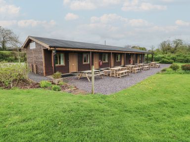 A single-story wooden building with multiple doors and windows next to picnic tables on a gravel area surrounded by grass at Oak in Cowbridge
