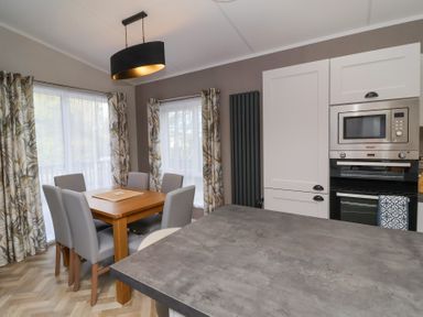 A dining area with a wooden table and six gray chairs next to windows with patterned curtains and a kitchen with white cabinets and built-in microwave at 16 Manleigh Park in Combe Martin