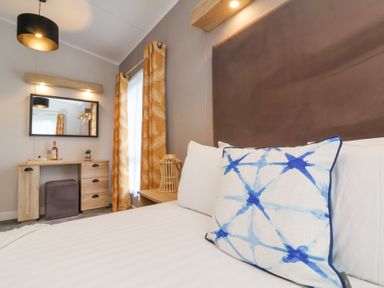 A bedroom with a bed with patterned pillows a wooden desk a mirror curtain and a ceiling lamp at 16 Manleigh Park Combe Martin
