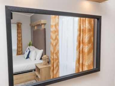 A reflection of a bedroom with a bed white pillows wooden nightstand and orange curtains with feather patterns at 16 Manleigh Park in Combe Martin