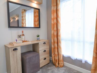 A wooden dressing table with a mirror above it and a gray ottoman underneath next to a window with orange curtains and white sheer curtains at 16 Manleigh Park in Combe Martin