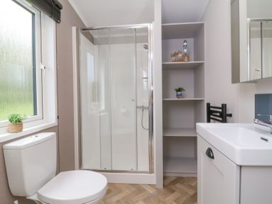 A bathroom with a toilet beside a window a glass shower enclosure open shelves with decorations and a sink cabinet at 16 Manleigh Park in Combe Martin