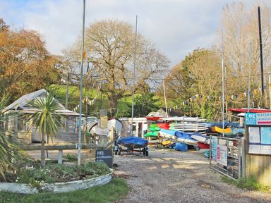 An outdoor boat and kayak hire area with boats and kayaks stacked near a wooden building at Loe Beach in Cornwall