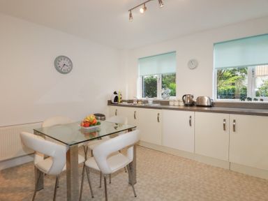 A kitchen with a glass dining table and four white chairs next to cream cabinets and windows with blue blinds at 13 Clock Tower Court in Charlestown