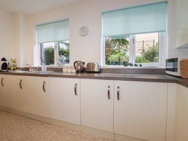 A kitchen with white cabinets a sink kettle toaster microwave and two windows with blue blinds at 13 Clock Tower Court in Charlestown
