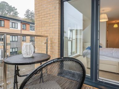 A balcony with a black metal table and chair overlooking apartments and a bedroom visible through glass doors at 13 Clock Tower Court in Charlestown