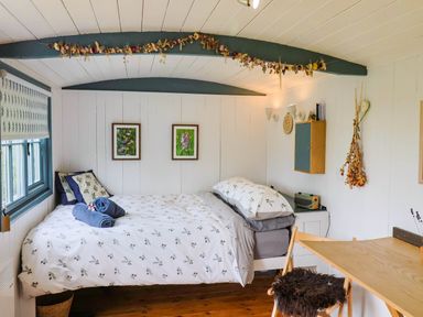 A bedroom with a bed with patterned bedding next to a window and a wooden desk with a chair at Oak Shepherd Hut Monkton Wyld near Charmouth