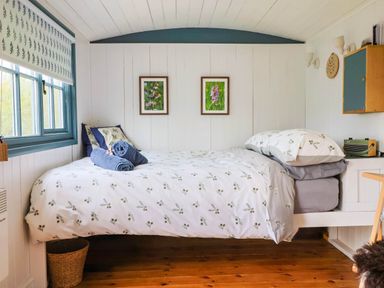 A bedroom with a bed, window with patterned blind, two framed pictures, wooden floor and a small cabinet at Oak Shepherd Hut Monkton Wyld near Charmouth