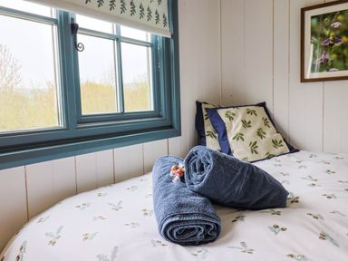 A bedroom with a window blue frame two rolled blue towels on a bed with leaf patterned bedding and matching cushions at Oak Shepherd Hut in Monkton Wyld near Charmouth