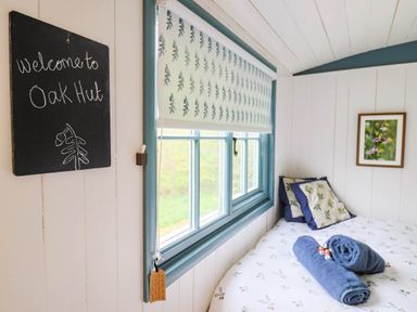 A bedroom with a bed cushions rolled towels window blinds and a welcome sign at Oak Shepherd Hut Monkton Wyld near Charmouth