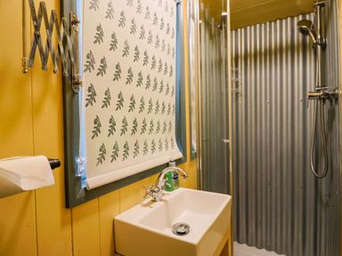 A small bathroom with a sink a window with a patterned blind a shower with corrugated metal walls and a toilet paper holder at Oak Shepherd Hut in Monkton Wyld near Charmouth