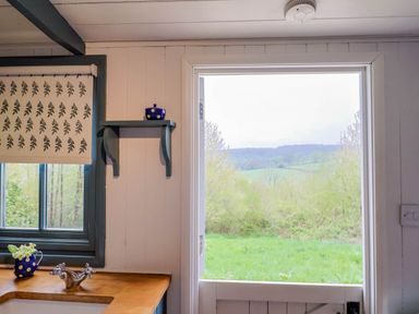 A kitchen countertop with a faucet and flower vase near a window and a door opening to green fields at Oak Shepherd Hut Monkton Wyld near Charmouth