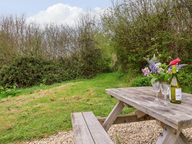 A wooden picnic table with a bottle of wine glasses and flowers on gravel next to grassy path with trees at Oak Shepherd Hut in Monkton Wyld near Charmouth