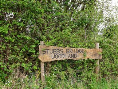 A wooden sign saying Stubbs Bridge Woodland surrounded by green bushes and grass