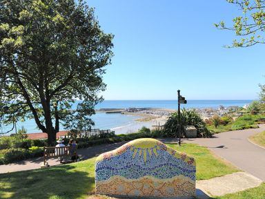A park with a mosaic bench and people sitting on another bench overlooking a beach and harbor
