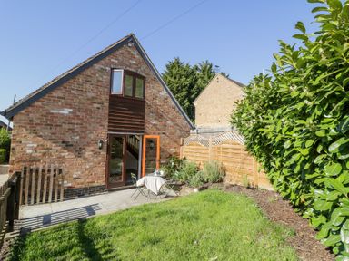 A brick house with an open door leading to a patio with a table and chairs next to a garden with green grass and shrubs at Little Barn in Dodwell near Stratford-Upon-Avon