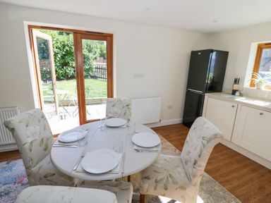 A dining area with a round table set for four and floral upholstered chairs near a kitchen with a black fridge and white cabinets at Little Barn in Dodwell near Stratford-Upon-Avon