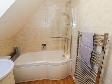 A bathroom with a bathtub shower combination, glass shower screen, towel radiator with towels, and beige tiled walls at Little Barn in Dodwell near Stratford-Upon-Avon