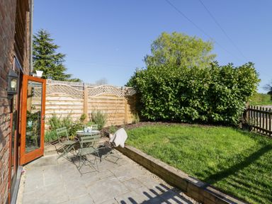 A garden with a small table and chairs on a paved patio next to a grassy area with bushes and a wooden fence at Little Barn in Dodwell near Stratford-Upon-Avon