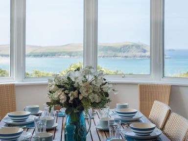 A dining table set with blue and white dishes and glasses with a floral centerpiece by windows overlooking the sea at Splits in Daymer Bay