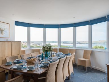 A dining room with a wooden table set with dishes and wicker chairs by large windows overlooking water at Splits in Daymer Bay