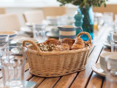 A basket with croissants and flowers on a wooden table set with cups and glasses at Splits in Daymer Bay