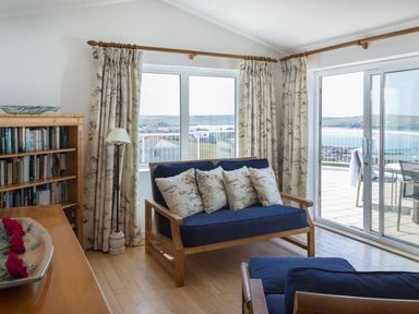 A living room with blue sofas, floral curtains, a wooden bookshelf, and sliding glass doors with a view of the sea at Splits in Daymer Bay