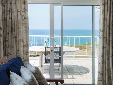 A balcony with a round table and chairs overlooking the sea seen through sliding glass doors with a sofa and curtains inside at Splits in Daymer Bay