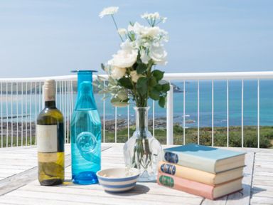 A table with a bottle of wine a blue bottle a bowl flowers in a vase and three books with a sea view at Splits in Daymer Bay