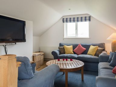 A living room with blue sofas yellow and red cushions a round wooden coffee table with red flowers a wall-mounted television and wicker baskets at Splits in Daymer Bay