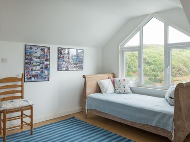 A bedroom with a wooden single bed with pillows near a large window and a wooden chair with a cushion on a striped rug at Splits in Daymer Bay