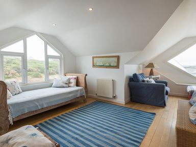 A room with a daybed beside a window an armchair a wicker sofa a blue striped rug and a wall painting at Splits in Daymer Bay
