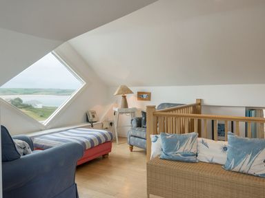 A sitting area with blue upholstered chairs a striped ottoman a lamp on a white table and a large diamond-shaped window at Splits in Daymer Bay