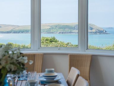 A dining area with a table set for a meal and woven chairs facing a window with a view of the sea and a coastal landscape at Splits in Daymer Bay
