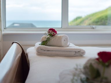A bed with folded white towels and a red flower on it near a window with a view of the sea at Splits in Daymer Bay
