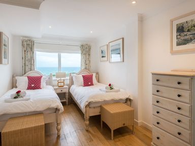 A bedroom with two single beds with red pillows a wooden dresser and a window showing a sea view at Splits in Daymer Bay