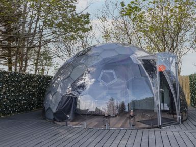 A transparent geodesic dome structure on a wooden deck surrounded by trees and hedges at Number 1 Dome in Derrybeg County Donegal