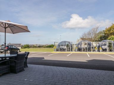 An outdoor seating area with tables chairs and umbrella near clear dome-shaped enclosures in a parking lot at Number 1 Dome in Derrybeg County Donegal