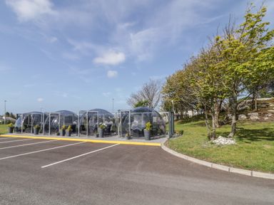 A row of transparent outdoor domes with furniture inside next to a parking lot and trees at Number 1 Dome in Derrybeg County Donegal