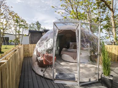A transparent geodesic dome with a bed and chairs inside on a wooden deck surrounded by a wooden fence at Number 2 Dome in Derrybeg County Donegal